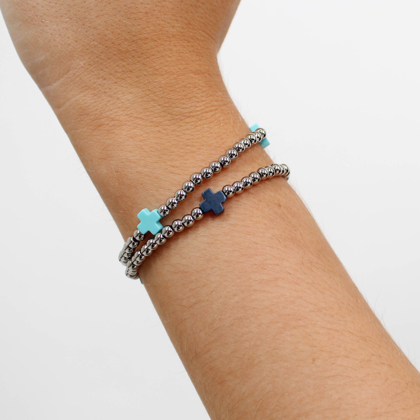 Two beaded silver cross bracelets in blue on a wrist against and white background. 