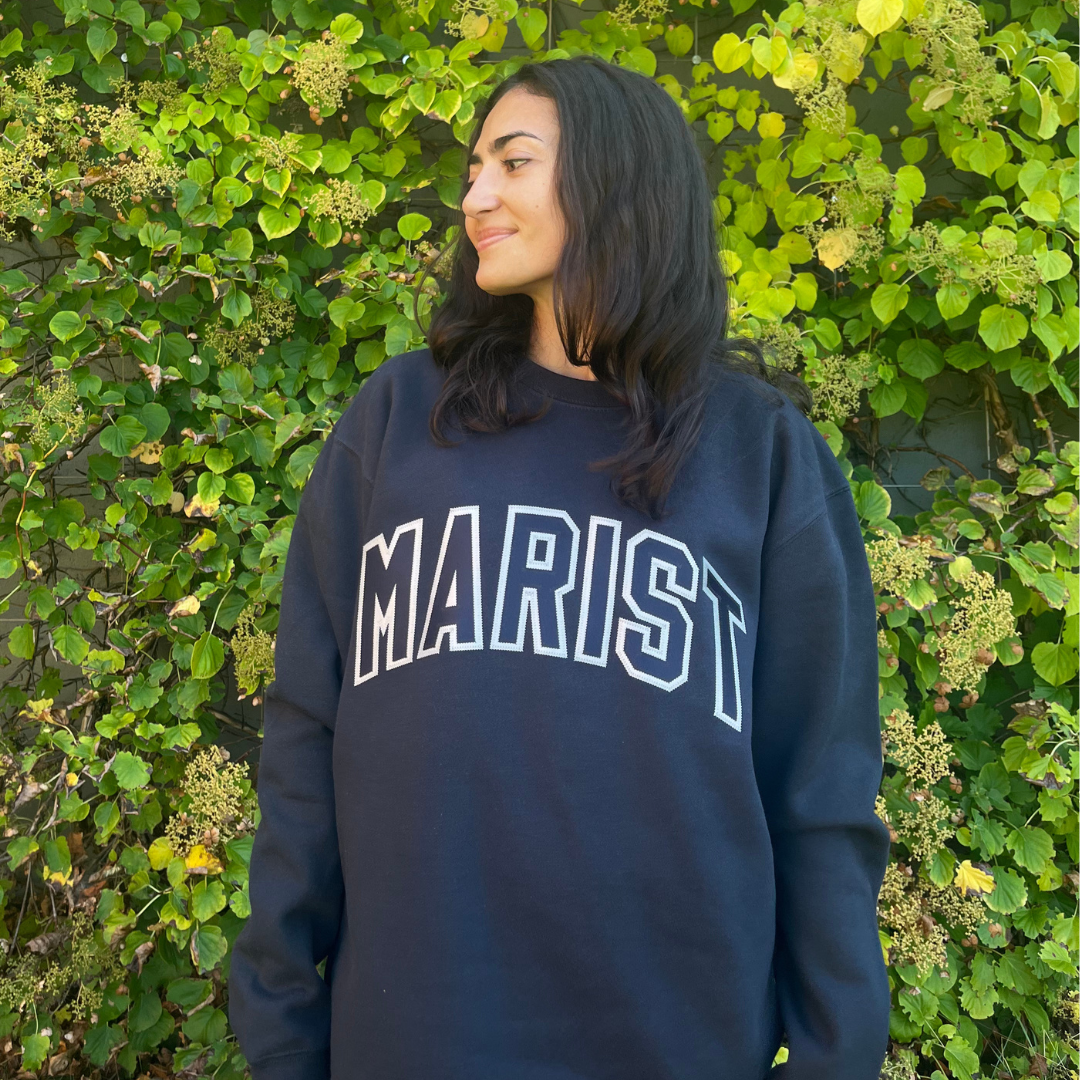 Person wearing a navy blue and grey 'MARIST' varsity crewneck sweatshirt standing against a green leafy background