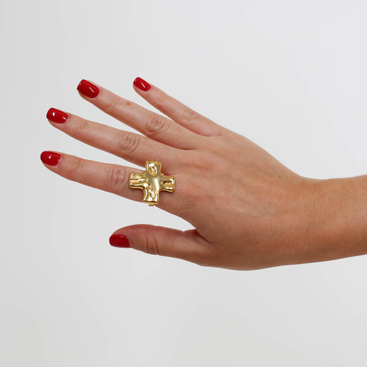 Hand wearing a gold statement cross ring on a plain background